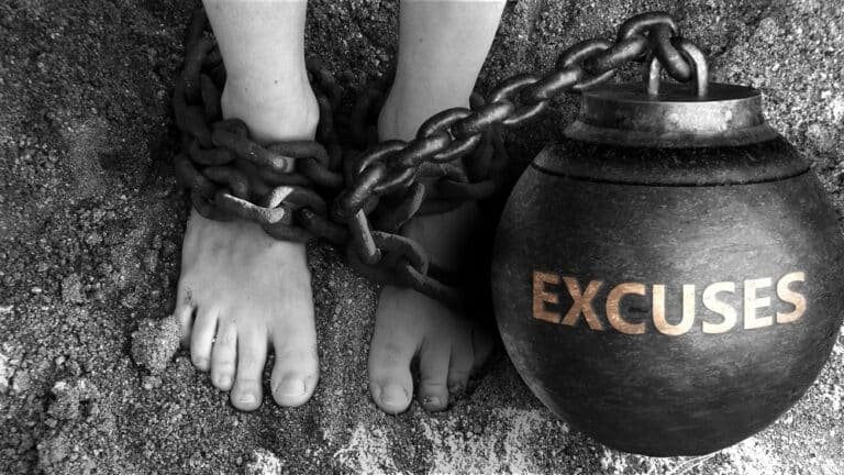 Pieds nus enchaînés à un lourd boulet noir marqué "EXCUSES", en noir et blanc, vue plongeante sur le sol