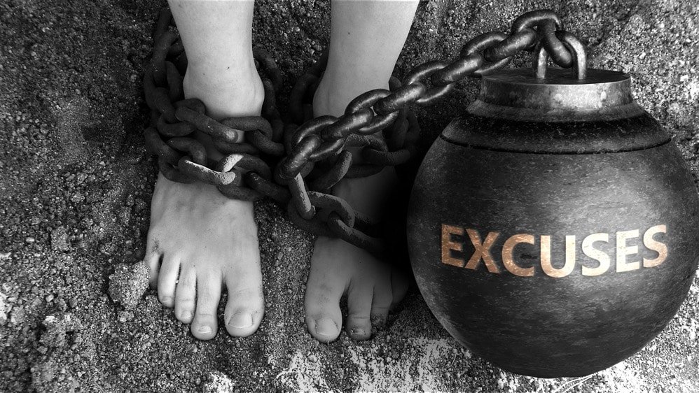 Pieds nus enchaînés à un lourd boulet noir marqué "EXCUSES", en noir et blanc, vue plongeante sur le sol